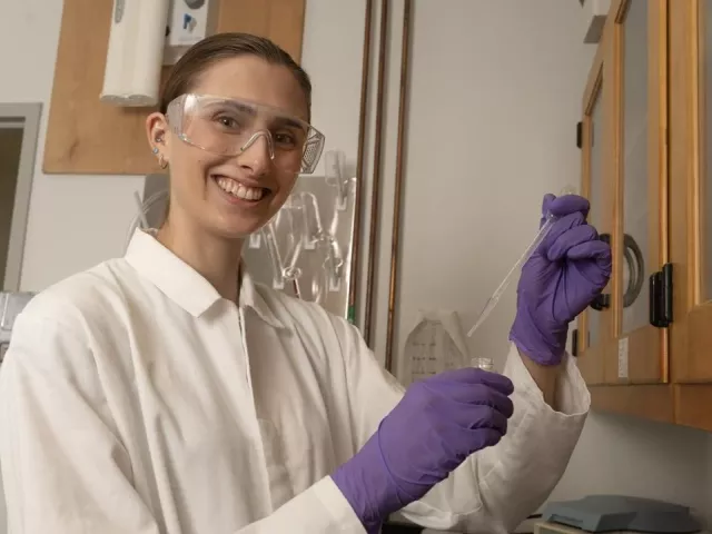 A woman wearing a lab coat and purple lab gloves working in a lab, smiling