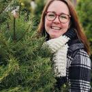 A woman wearing glasses smiling at the camera near a pine tree.